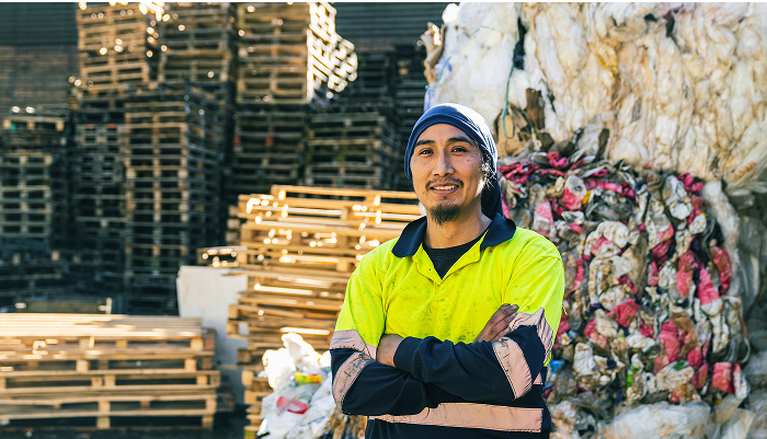 Man standing with his arms crossed, piles of garbage and pallets behind him